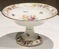 Clear photo showing porcelain pedestal pastry plate with floral design and gold trim, highlighting its shape and decoration.