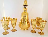 Photo showing 1 decanter with stopper and 6 matching cups arranged in a row, all gold glass with black classical scene designs.