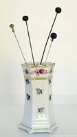 Front view of the hexagonal white porcelain hatpin holder with colorful floral decoration and gold trim, with 4 hatpins inserted.