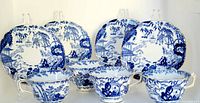 Four blue and white oriental pattern tea cups displayed with four matching saucers behind them. Pattern shows pagodas, bridges, trees, and figures. Cups and saucers are square-shaped with scalloped edges on saucers and gold trim on cups.