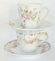 Two vintage Limoges demitasse cups with matching saucers featuring floral patterns and gold accents, stacked one atop another. The larger cup and saucer have pink floral designs; the smaller set has pastel pink and blue flowers with gold detailing.