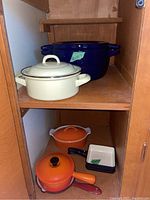 Complete shelf with orange Le Creuset pot with lid, red Dansk pan, small white enamel dish with handle, orange enamel saucepan with handle
