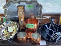 Photo of large etched serving platter, copper pot, copper cups, silver-tone serving bowl with utensils, and black cast iron pan with kitchen utensils.
