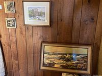 Four framed artworks on wood-paneled wall including large seascape painting, 'River Rapids - Elora' print, and two small native art plaques