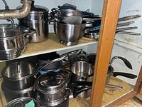 Side view of shelf with multiple pots, pans, and colander, showing metal and non-stick cookware with handles and lids.