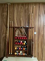 Full view of the vintage wooden cue rack holding multiple wooden pool cues on the sides, standard billiard balls, red cue balls on shelves, and a metal triangular ball rack attached in the center.