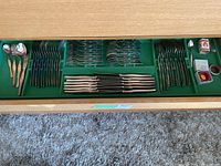 Full view of silverware set in green felt-lined drawer showing knives, forks, spoons, and wine collars, along with small boxes.