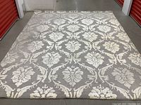 Top-down photo showing full 8x10 grey area rug with off-white floral medallion pattern spread flat on concrete floor. Visible staining present.