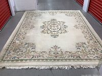 Full view of the rug showing overall design, cream base, floral medallion in center, wide floral border and fringed edges. Visible discoloration spots.