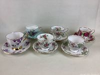 Full front view of all six cups and saucers arranged in a row showing floral patterns and gold accents on fine china.
