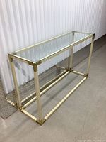 Console table with clear glass top and shiny brass accented metal frame, photographed against a white paneled wall and concrete floor.
