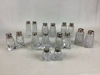 Front view of eight pairs of clear vintage crystal salt and pepper shakers in varied cut patterns with silver-plated tops arranged in two rows on a white background.
