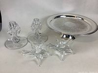 Photo showing silver pedestal cake tray engraved with floral patterns, two ornate glass candlesticks, and two flower-shaped glass bud vases.