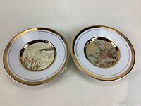 Two decorative ceramic plates with white base, gold rim, and detailed center designs with cranes and floral patterns.