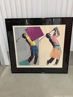 Framed abstract art print depicting two male golfers in mid-swing, using a Warhol-style pop art technique with bright colors and large colored blocks in the background. The frame is black with glass and the print is matted with a cream-colored border.