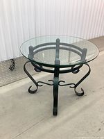 Side angle of the round glass top table with visible circular metal frame and curved legs on light concrete floor.