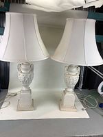 Pair of full lamps with white shades shown from front and angled side angle. Both lamps have Grecian style white ceramic bases and matching white shades.