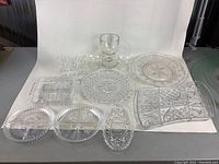 Top-down view of assorted clear glassware on white backdrop showing plates, trays, and stemware including textured and pressed glass patterns.