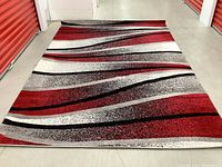 View of the modern area rug laid out in a storage unit, showing the full pattern with red, white, black and gray wavy stripes.