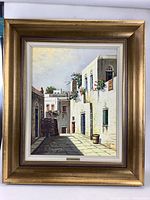 Frontal view of the signed oil painting in a gold frame showing a sunlit alleyway with white buildings and vibrant flowers.