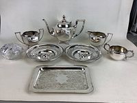 Complete set showing all items including the tea pot, cream and sugar containers, two decorative silver bowls, two plates, cut glass bowl, thimbles, and serving tray.