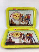 Two yellow bordered vintage Coca-Cola metal trays with designs of people, a dog, and Coca-Cola Classic logos dated April 21, 1980