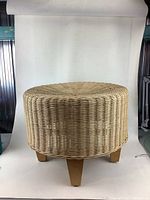 Round wicker foot stool photographed from the front against a white backdrop, showing its woven wicker texture and wooden legs.