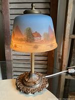 Full view of the antique lamp showing the painted glass shade and ornate metal base on a glass table.