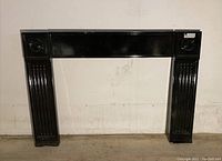 Full frontal image of the gloss black metal fireplace piece showing its rectangular shape and vertical columns with decorative elements.