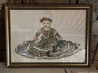 Full view of the large needlepoint artwork showing a seated girl in a detailed dress and bonnet, mounted in an ornate gold-colored frame.
