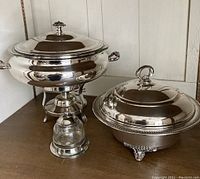 Photo showing a large silver plate covered server with stand and another large silver plate server with a lid, both placed on a wooden surface.