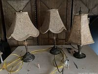 Three table lamps on a table: two with scalloped beige fabric lampshades and one with a square tapered fabric lampshade. All bases are dark metal and decorative.