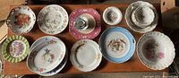 Top-down photo showing a collection of ten decorative dishes and plates arranged in three rows on a wooden surface. Plates show various floral and fruit patterns with distinctive scalloped, pierced, and curved edges.