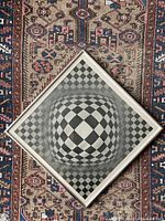 Top down photo showing complete square metal framed print on patterned carpet background, highlighting the geometric diamond checkerboard pattern creating a 3D bulging effect.
