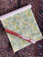 Two rolls of wallpaper with floral pattern on a carpeted surface, one roll partially unrolled showing yellow and green flowers on white background.