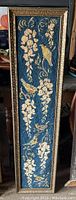 Full view of the tall rectangular framed needlepoint artwork showing birds and cascading flowers on a blue background.