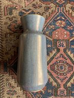 Full view of the Bay Keramik vase showing its metallic-toned cylindrical body with slight flare at the top, resting on a patterned rug.