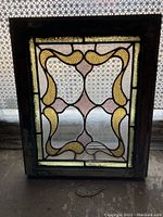 Full view of vintage stained glass window with floral and geometric pattern, shown against a patterned background, exhibiting some damage and wear to frame and glass.