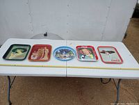 Five Coca-Cola collector tin trays displayed on a white table, showing full lot.