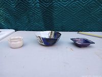 Photo of three decorative pottery pieces on a table: a white scalloped bowl, a hexagonal multi-color glazed bowl, and a small rectangular purple dish.