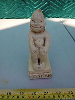 Front full view of aged good luck statue showing whole figure and base with inscription, placed beside ruler for scale.