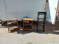 Three wooden furniture pieces outdoors including two small shelving carts on wheels and one tall black shelving unit with heart cut-out.