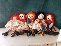 Group of eight Raggedy Ann and Andy dolls sitting on white surface with blue fabric background. Dolls have varied hair colors including red, orange, black yarn, and varied outfits with black shoes on some.