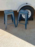 Two blue industrial metal stools standing on outdoor pavement, showing full view and relative size.