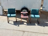 Front view of two wooden armchairs with blue patterned fabric cushions and a wooden side table between them, outdoors in sunlight.