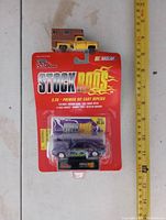 Photo showing unopened Stock Rods NASCAR diecast car in original packaging next to vintage yellow and brown toy truck and a measuring tape for scale.