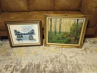 Pair of framed landscape paintings, winter scene and forest river scene on floor against couch