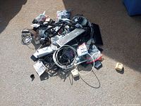 Photo of a large pile of mixed electronic items including cables, a wired keyboard, power adapters, and various small devices scattered on the ground.