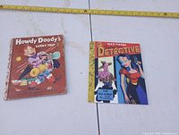 Two kids books shown side by side, a worn hardcover 'Howdy Doody's Lucky Trip' and a colorful softcover 'Max Hamm Fairy Tale Detective Vol. 1'. A measuring tape above shows approximate width of books.
