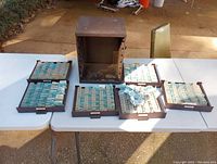 Full view of vintage rusty metal cabinet with five trays arranged on a white table, each tray filled with replacement watch crystals in paper packages.